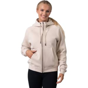A women's cotton zippered hoodie in beige with a kangaroo pocket and a logo. Model is smiling and wearing black leggings.