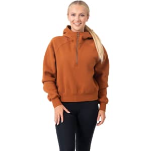 A woman wearing an orange half-zip jumper with a hood, paired with black leggings. The jumper has a logo.