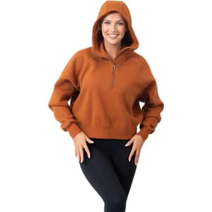 A woman poses in a rust-coloured half-zip hoodie with a hood, paired with black leggings. The hoodie features a small logo.