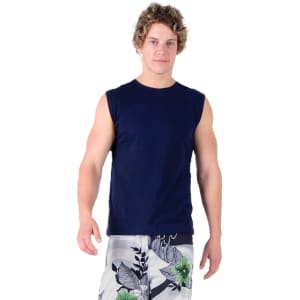 A man in a navy muscle T-shirt and patterned shorts stands on green grass, with trees in the background.
