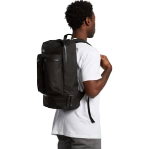 A person wearing a black recycled travel backpack, featuring multiple pockets and a logo on the front.