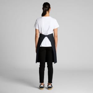 A person wearing a black Carrie Half Apron over a white T-shirt and black pants, facing away from the camera.