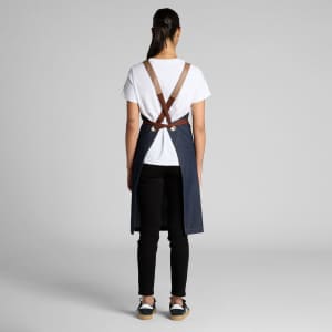 A denim apron with brown straps, worn by a person facing away, paired with a white t-shirt and black trousers.