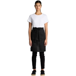 A model wearing a black linen half apron over a white t-shirt and black pants, standing straight with hands at her sides.