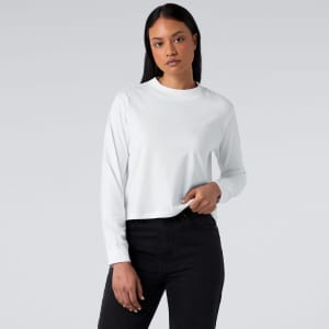 A woman wearing a white long-sleeve T-shirt, with dark jeans and a neutral background. The shirt features a logo.