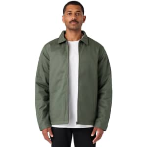 A man wearing a green service jacket over a white t-shirt, standing with a neutral expression. The jacket has a logo.