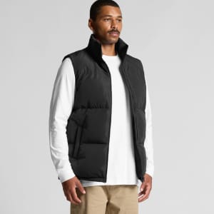 A man in a black puffer vest over a white long-sleeve shirt, standing against a neutral background.