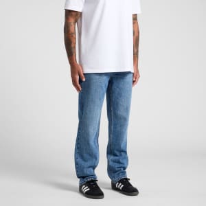 A pair of straight blue jeans worn by a person with tattoos, standing in a white t-shirt and black sneakers.