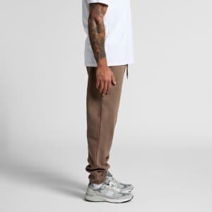 Brown track pants with a relaxed fit and an elastic waistband, paired with grey sneakers, showcased from the side.