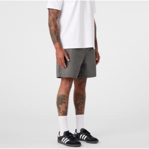 A man stands in grey dress shorts, paired with a white t-shirt and black sneakers, showing his tattoos.
