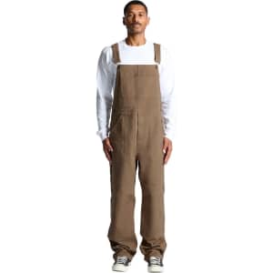 Canvas overalls in brown, featuring adjustable straps and a relaxed fit. Model is wearing them with a white long-sleeve shirt.