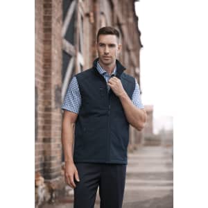 A man in a navy vest over a checked shirt stands confidently in an industrial setting. The vest features a logo.
