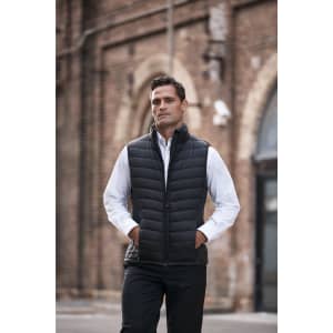 A man in a black puffer vest stands with hands in pockets, wearing a white shirt against a brick wall backdrop.