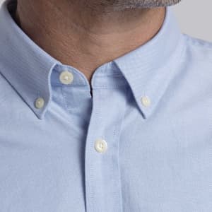 A close-up of a light blue button-up shirt featuring a classic collar and white buttons, with a branding logo present.