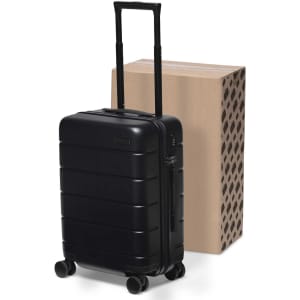 A black hardcase trolley with four wheels and a retractable handle, positioned next to a cardboard box.