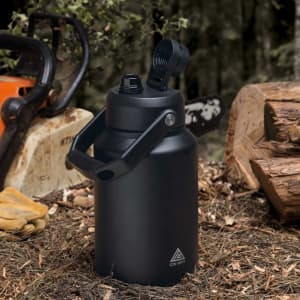 A drink bottle in matte black with a handle and a flip-top spout, set on a forest floor. It features a logo.