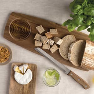 A wooden artisan tasting board with bread, cheese, honey, and a knife, surrounded by basil and a drink.