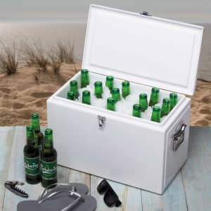 A white cooler box filled with green bottles of beer, sitting on a sandy beach with sunglasses and a bottle opener nearby.