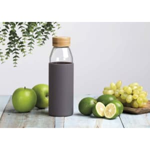 A drink bottle with a clear glass body, grey silicone sleeve, and a bamboo lid, surrounded by green apples and grapes.