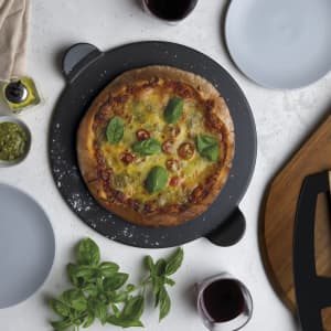 A pizza on a black serving stone, garnished with basil and cherry tomatoes, surrounded by plates and wine glasses.