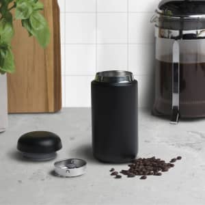 A sleek black drink bottle with a removable lid, placed beside coffee beans and a coffee maker on a countertop.