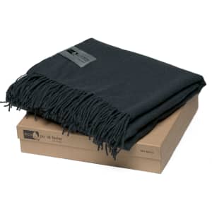 A dark green Merino blanket with fringed edges, neatly folded on a card box. Features branding.