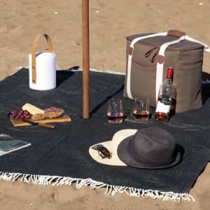 A dark blue picnic blanket on sand with a cooler bag, drink bottle, glasses, snacks, hat, and magazine.