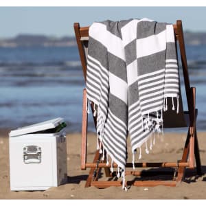 A grey and white striped Patara Turkish towel draped over a wooden beach chair beside a cooler on the sand.