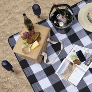 A picnic setup on a checkered rug featuring a timber picnic basket, wine, snacks, and a magazine.