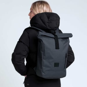 A black and grey roll-top backpack is worn by a person in a black jacket, featuring a logo on the backpack.