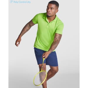 A man wearing a bright green polo shirt and navy shorts holds a badminton racket. The shirt features a logo.