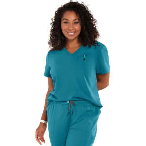 A woman in a teal scrub top with a v-neck and a chest pocket, smiling and standing with hands behind her back.