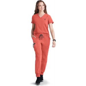 A woman in an orange scrub set walks confidently, featuring a V-neck top and drawstring pants. The outfit includes a logo.