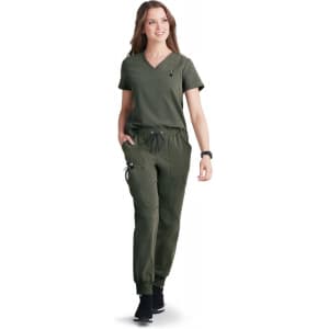A woman in an olive green scrub top and matching pants, walking confidently and smiling. The outfit has a logo.