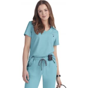 A woman models a teal scrub top with a v-neck and short sleeves. The outfit includes pockets and has a logo.