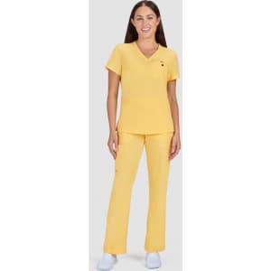 A woman stands in a yellow scrub set, featuring a short-sleeve top and matching trousers. The outfit has a logo.