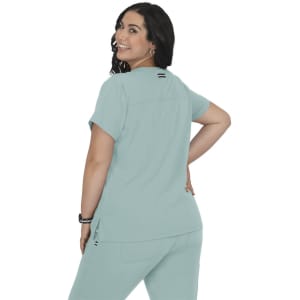 A woman wearing a mint green top with a relaxed fit, featuring a logo on the back, smiling and looking over her shoulder.