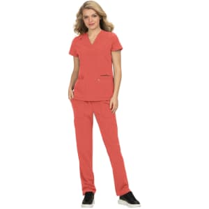 A woman wears a coral-coloured scrub top with matching pants, featuring a logo. She stands in a confident pose.
