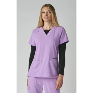 A lavender scrub top with short sleeves, featuring two front pockets. The model is wearing a black long-sleeve top underneath.