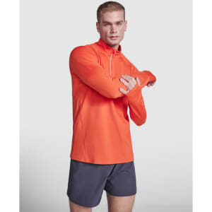 An orange half zip sweatshirt is worn by a model with short hair, paired with dark shorts. The garment includes a logo.