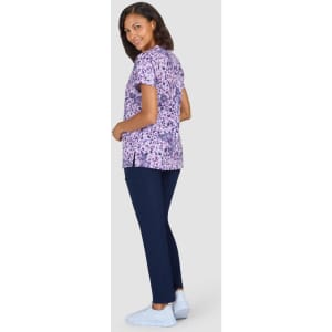 A woman wearing a purple patterned top and navy trousers stands facing sideways, smiling at the camera.