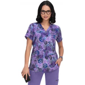 A woman wearing a purple floral print scrub top, smiling while holding a phone. The top has a logo.