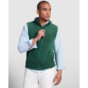 A green fleece vest with a high collar and front zip, modelled by a man in a light blue shirt and white pants.