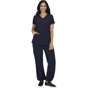 A woman stands wearing navy scrubs with a V-neck top and relaxed fit pants. The outfit includes pockets and has a logo.