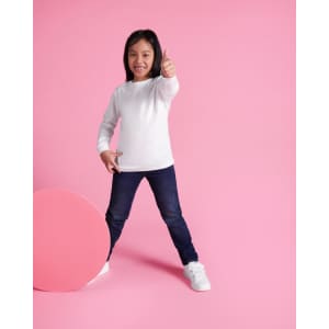 A child poses in a white long-sleeve t-shirt and blue jeans against a pink background, giving a thumbs-up.