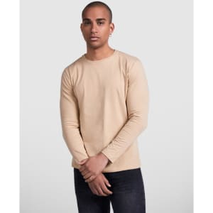 A man wearing a beige long-sleeved t-shirt, standing with arms crossed, against a neutral background.