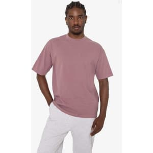 A man stands wearing a short sleeve, crewneck tee in a dusty pink colour, paired with grey bottoms. The tee has a logo.