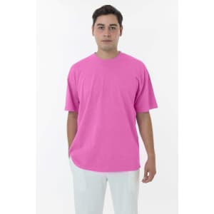 A man wears a bright pink short sleeve crewneck tee, paired with white trousers. The tee has a subtle logo.