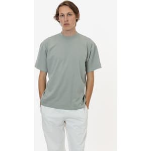 A short sleeve crewneck tee in muted green, featuring a relaxed fit and a logo. Styled with light-coloured trousers.