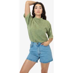 A model wearing a green mineral wash short sleeve crew tee paired with blue denim shorts, with a logo visible on the tee.